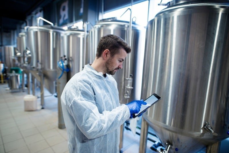 Supervisor de planta revisando un lote en una tablet junto a tanques de acero inoxidable